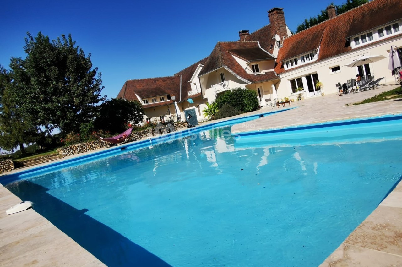 Piscine extérieure bordée de dalles, devant une grande maison traditionnelle avec toit en tuiles rouges.
