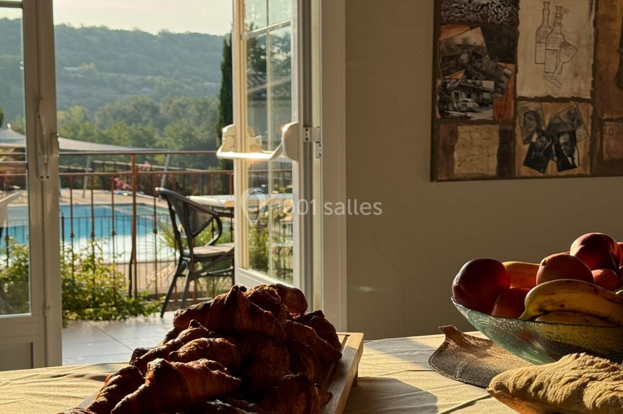 Vue d'une table avec croissants et fruits, devant une porte ouverte sur une terrasse avec piscine et paysage verdoyant.