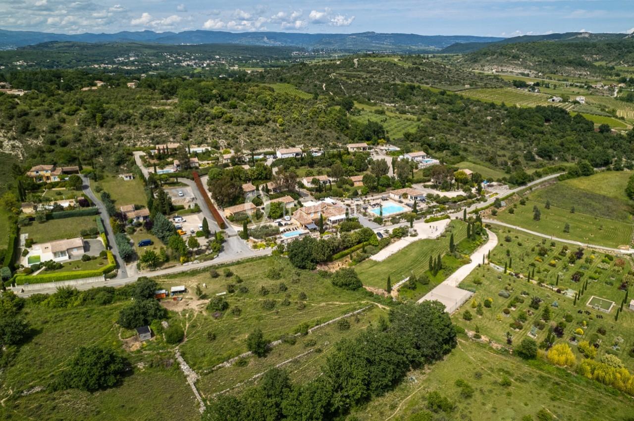 Vue aérienne d'un domaine avec bâtiments, piscine, jardins et paysages vallonnés environnants sous un ciel dégagé.