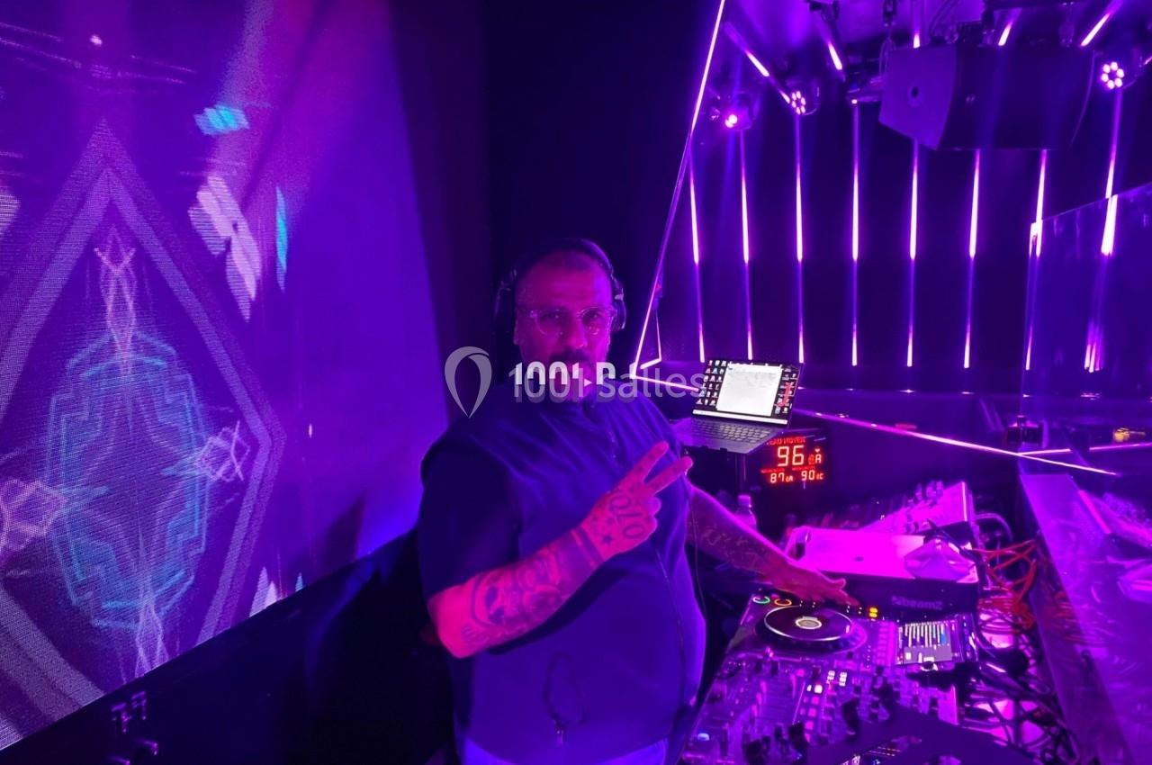 Un DJ mixe sur une table de mixage dans une ambiance lumineuse violette, faisant un signe de la main vers l'objectif.