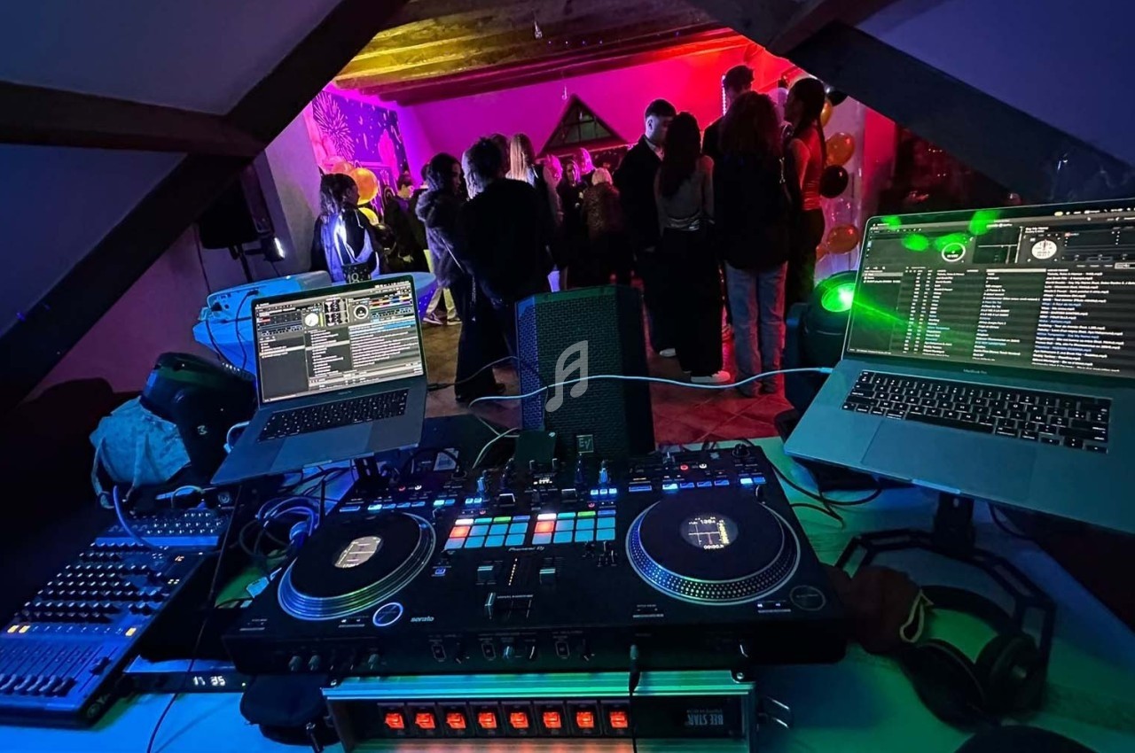 Vue d'une table de mixage avec deux ordinateurs portables, face à une fête dans une pièce éclairée par des lumières colorées.