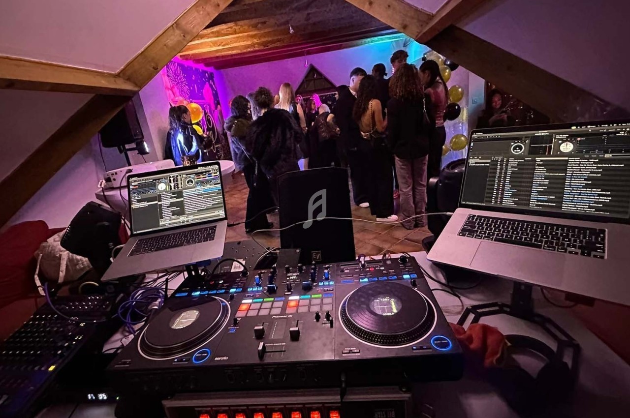 Vue depuis une table de mixage avec deux ordinateurs, face à un groupe de personnes dansant dans une pièce éclairée.