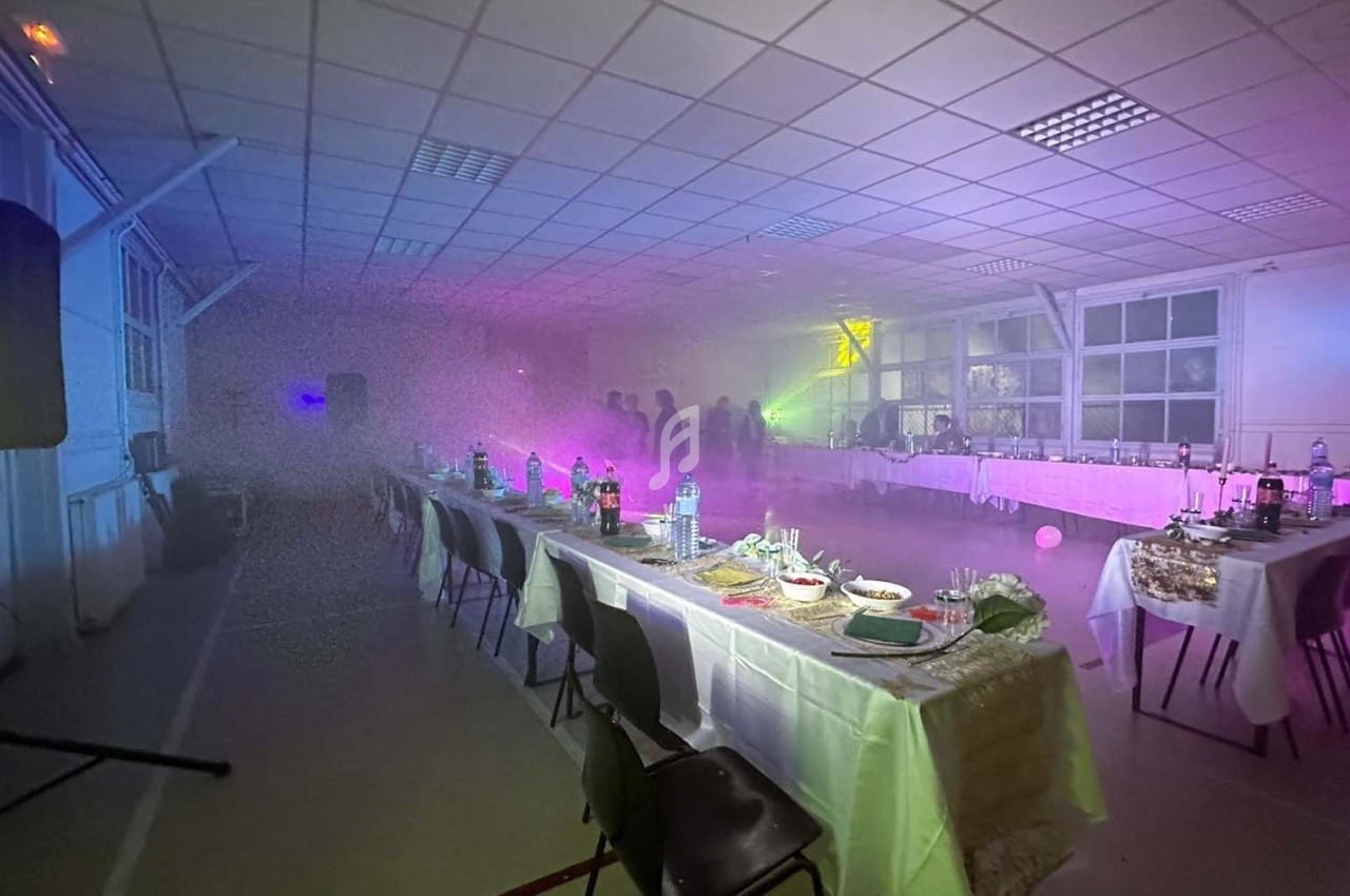 Salle de réception avec tables dressées, éclairage coloré et ambiance enfumée, silhouettes en arrière-plan.