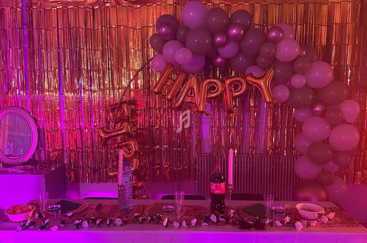 Décoration festive avec ballons, guirlandes dorées et table dressée contenant boissons, bougies et collations.