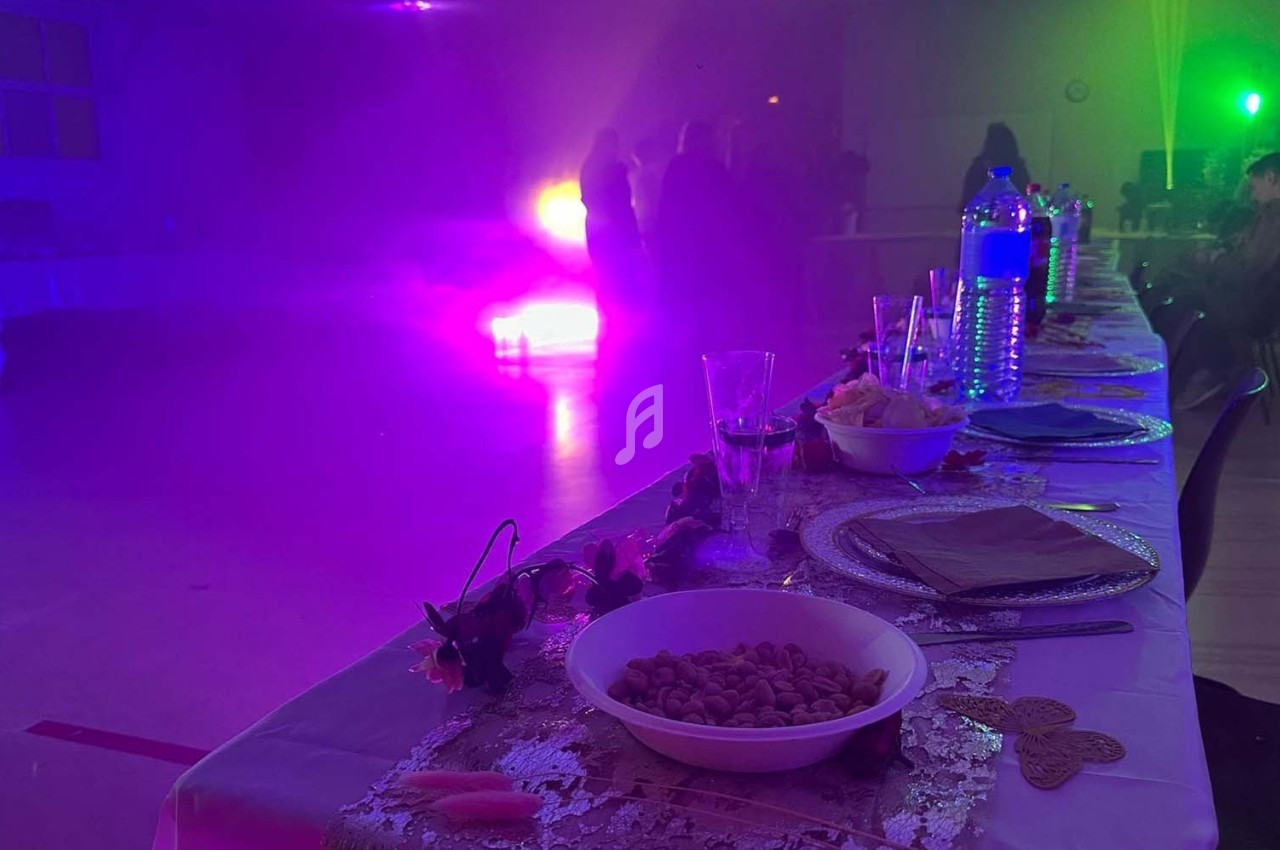 Table dressée avec des plats et boissons au premier plan, éclairage coloré et silhouettes de personnes en arrière-plan.
