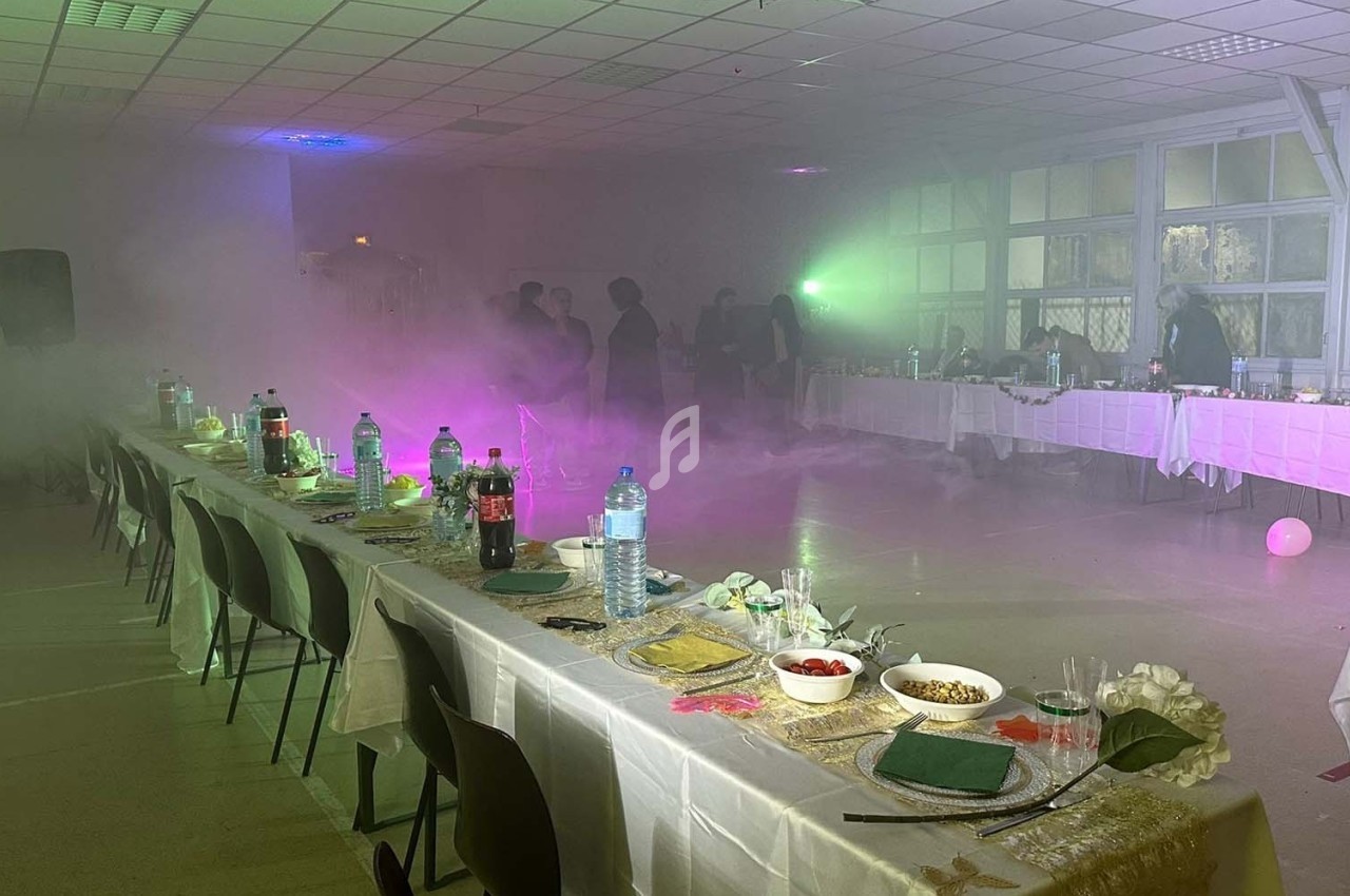Salle de réception avec tables dressées, éclairage tamisé et fumée diffuse, quelques personnes en arrière-plan.
