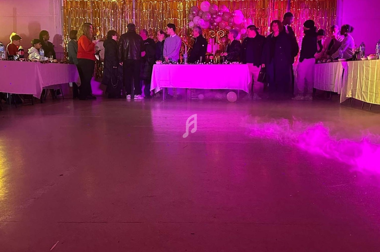 Des personnes rassemblées dans une salle décorée avec des ballons et des guirlandes dorées, éclairée par des lumières…