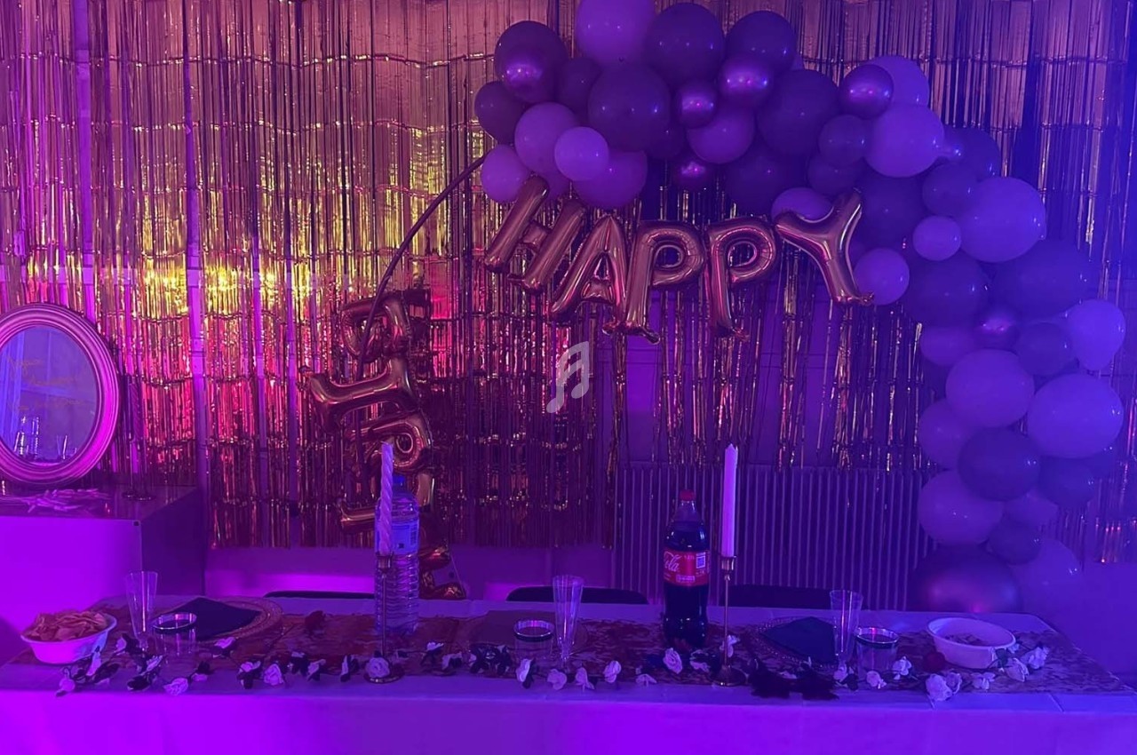 Décoration festive avec ballons violets et dorés, guirlande ’Happy’, table dressée et fond doré scintillant.