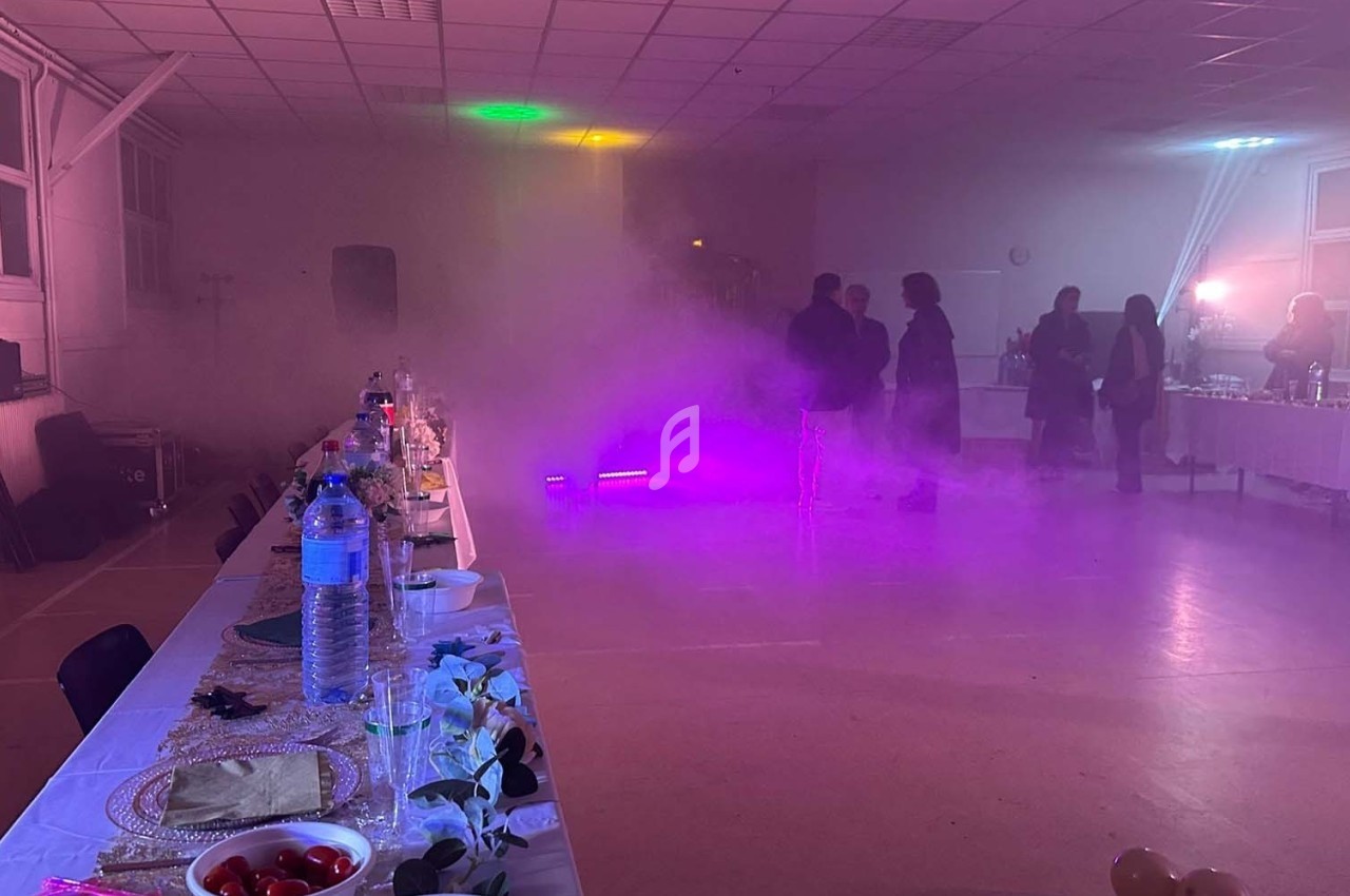 Salle de réception avec tables dressées, éclairage coloré et fumée légère, quelques personnes en arrière-plan.