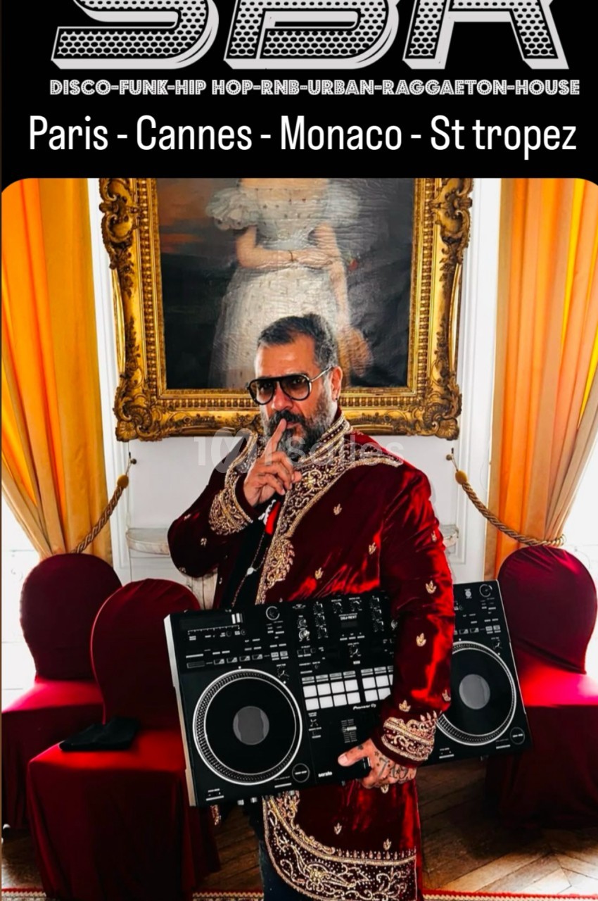Un homme en tenue élégante tient un micro et une table de mixage dans une pièce ornée de rideaux et d'un tableau.