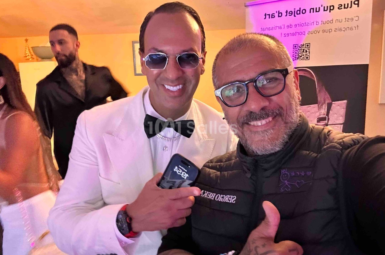 Deux hommes souriants posent pour une photo dans un environnement intérieur, avec d'autres personnes en arrière-plan.