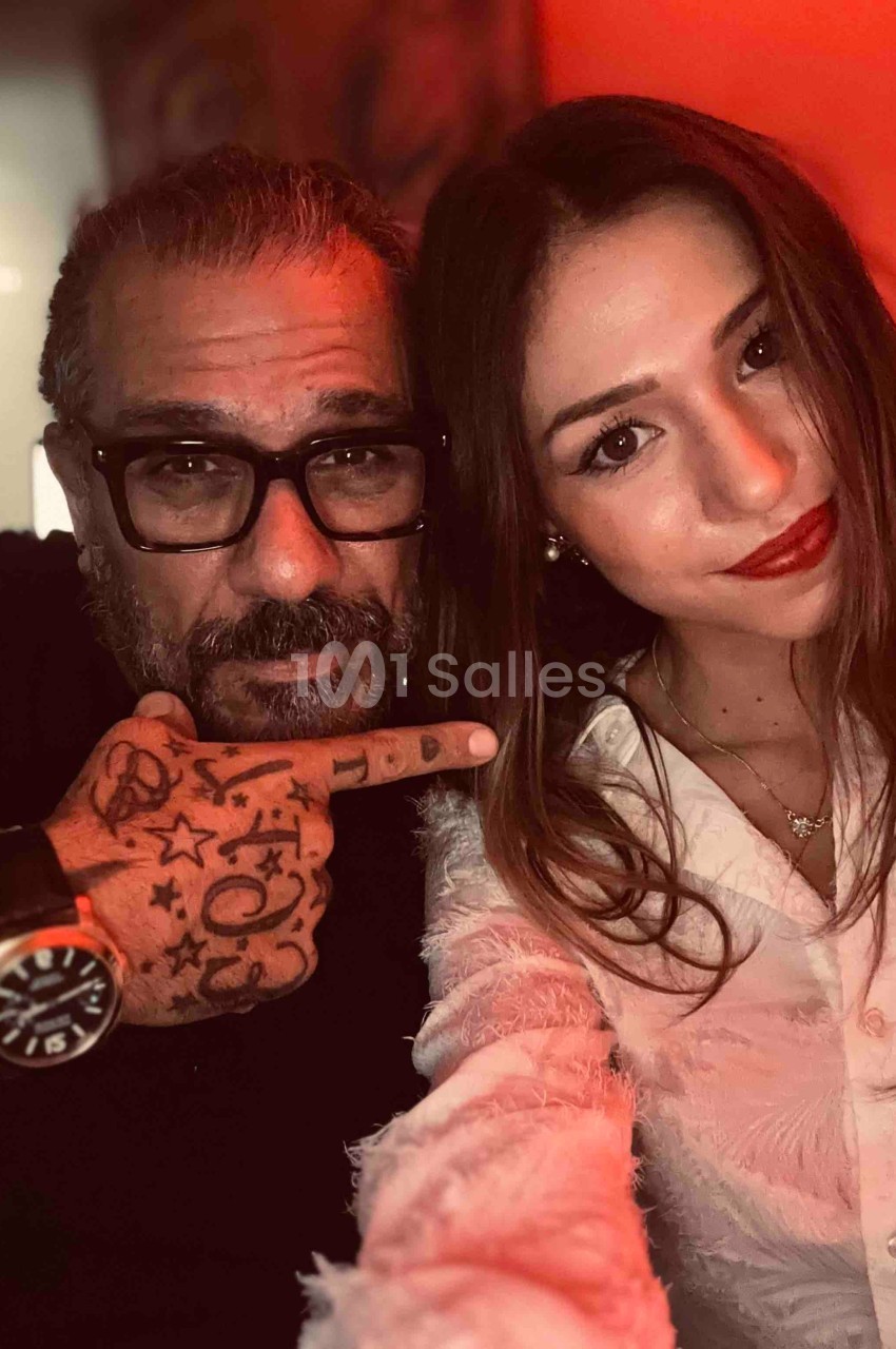 Un homme barbu avec des lunettes et des tatouages pose à côté d'une femme souriante dans un éclairage rouge tamisé.
