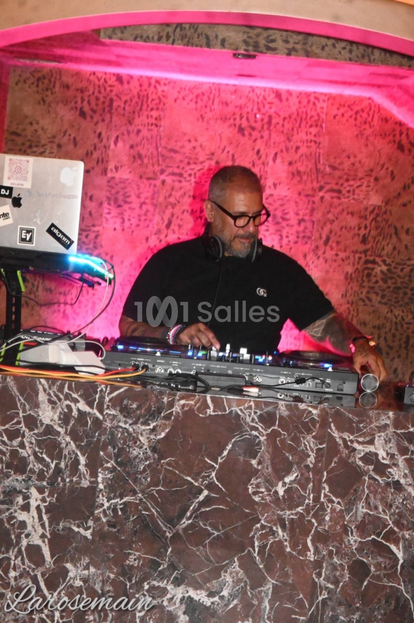 DJ en cabine équipée de platines, éclairage rose et fond texturé.