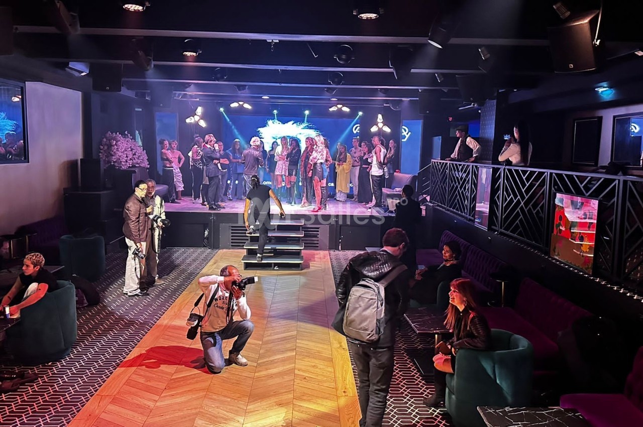 Salle de spectacle avec des personnes sur scène et dans le public, ambiance animée avec éclairages colorés.