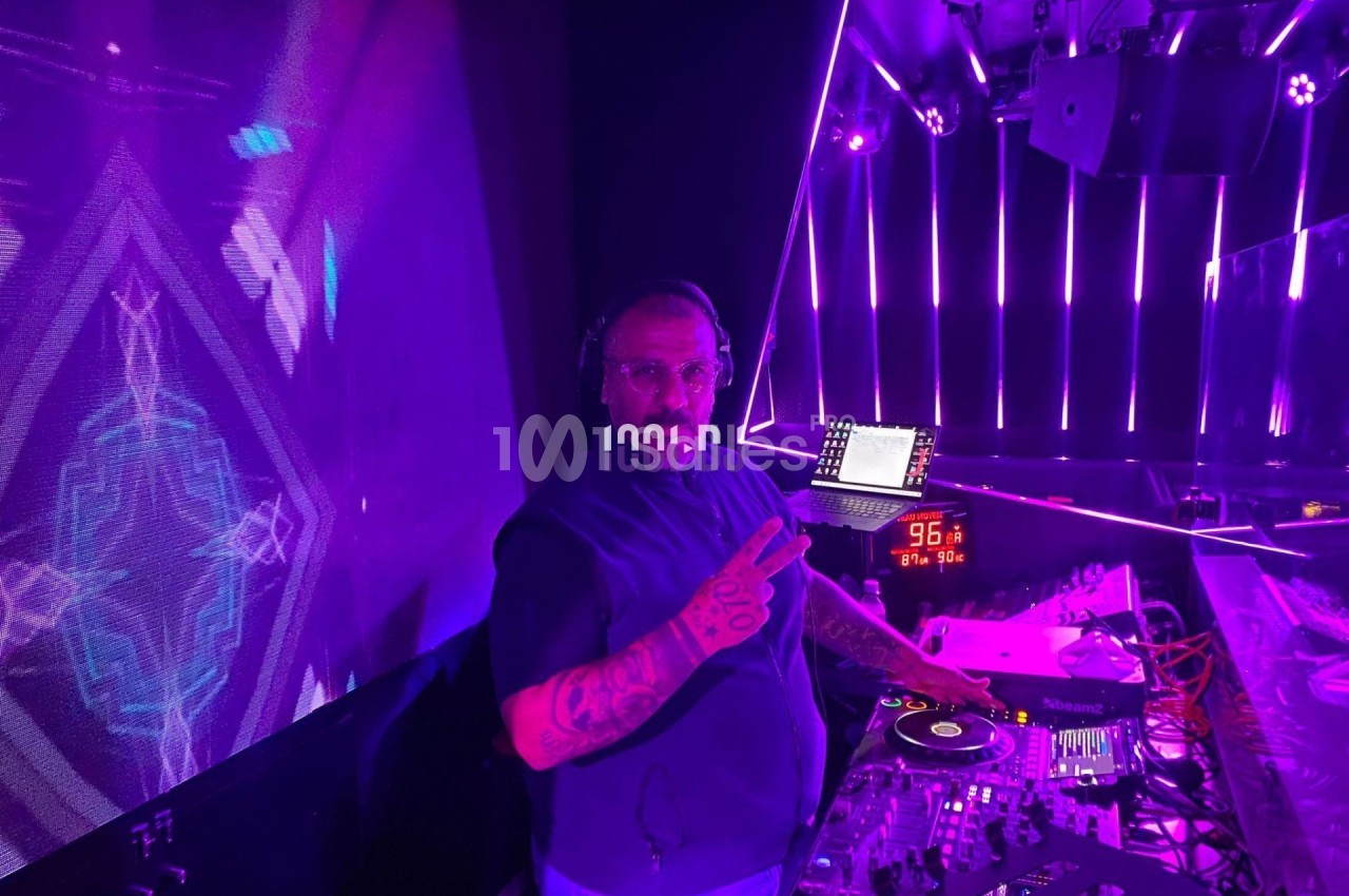 Un DJ mixe sur une table de mixage dans une ambiance lumineuse violette, faisant un signe de la main vers l'objectif.