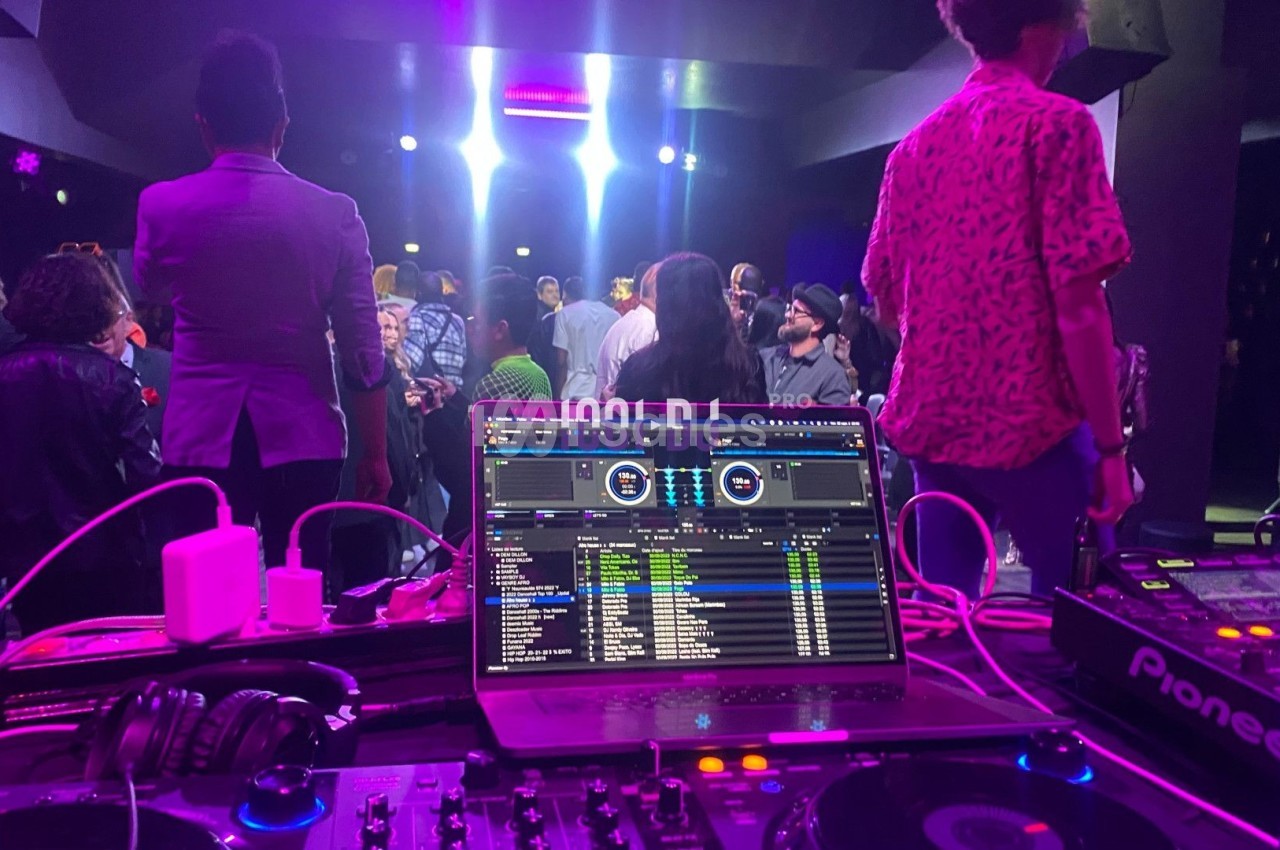 Vue depuis une table de mixage avec un écran affichant des pistes audio, face à une foule dans une salle éclairée.