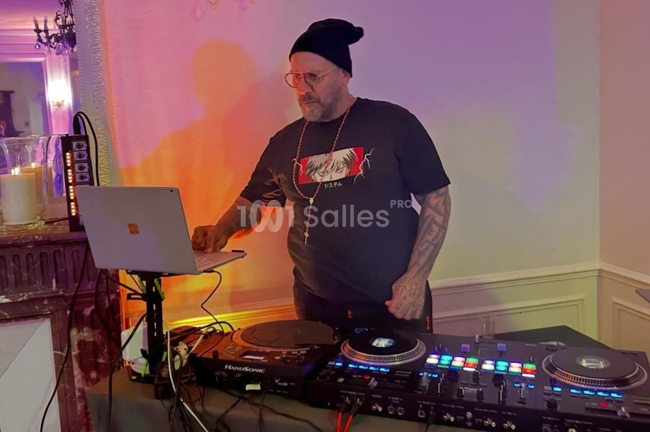Un DJ portant un bonnet mixe de la musique sur une console, avec un ordinateur portable à ses côtés dans une pièce éclairée.