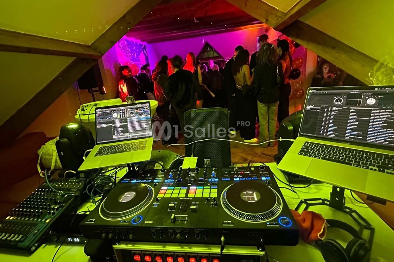 Vue d'une table de mixage avec ordinateurs et platines, face à une salle où des personnes dansent sous des lumières colorées.