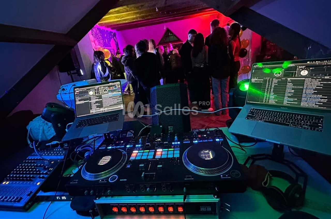 Vue d'une table de mixage avec deux ordinateurs portables, face à une fête dans une pièce éclairée par des lumières colorées.