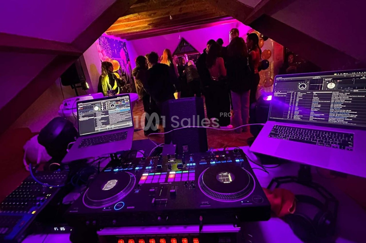 Vue d'une table de mixage avec deux ordinateurs, des platines et une fête en arrière-plan sous un éclairage coloré.