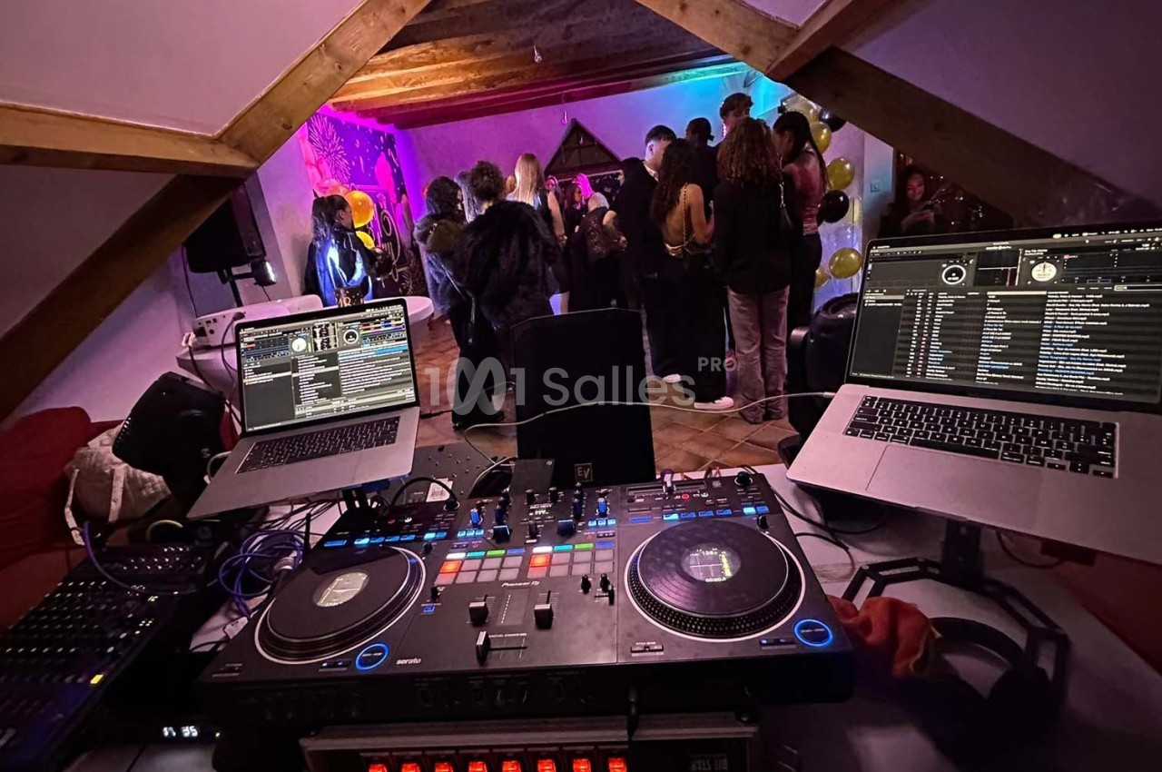 Vue depuis une table de mixage avec deux ordinateurs, face à un groupe de personnes dansant dans une pièce éclairée.