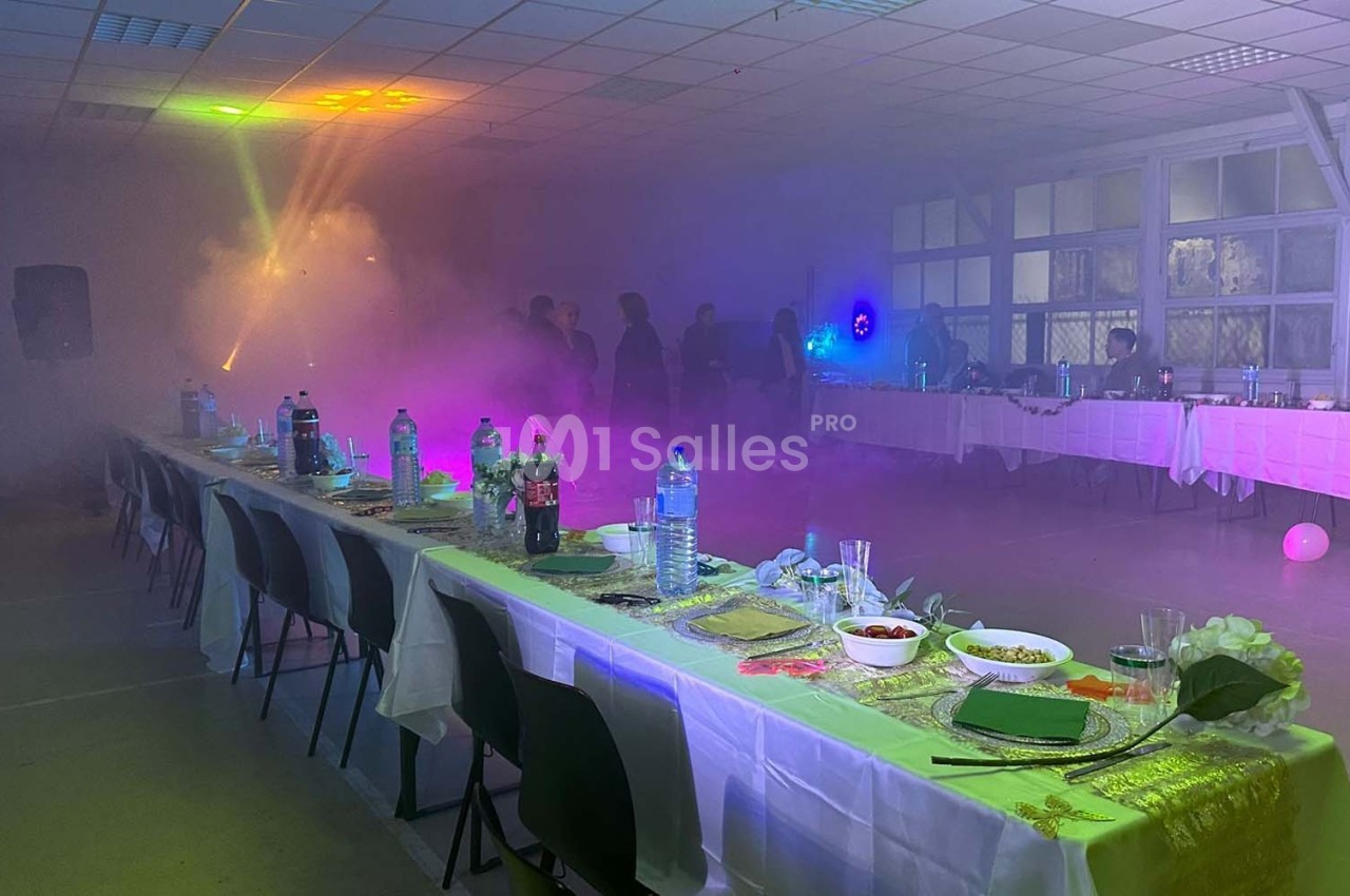 Salle décorée pour un événement avec tables dressées, éclairages colorés et ambiance enfumée.