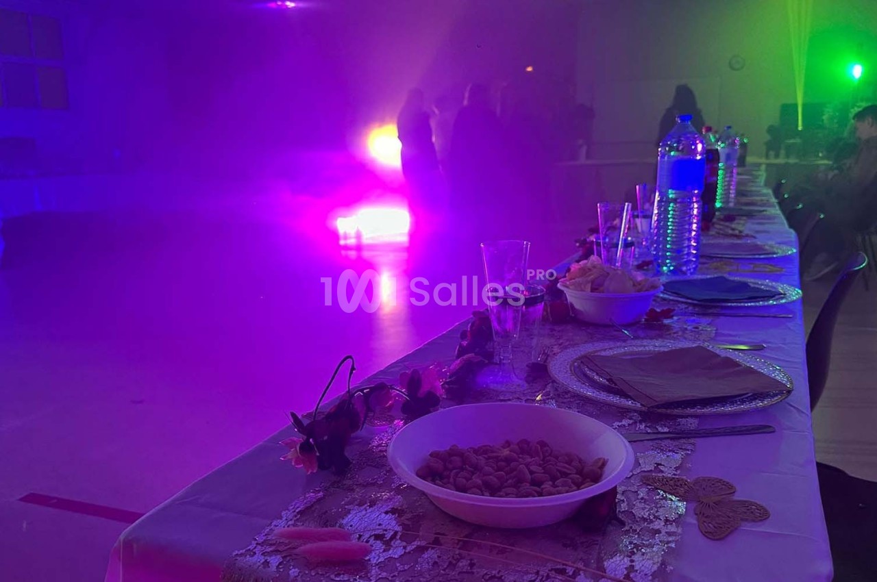 Table dressée avec des plats et boissons au premier plan, éclairage coloré et silhouettes de personnes en arrière-plan.