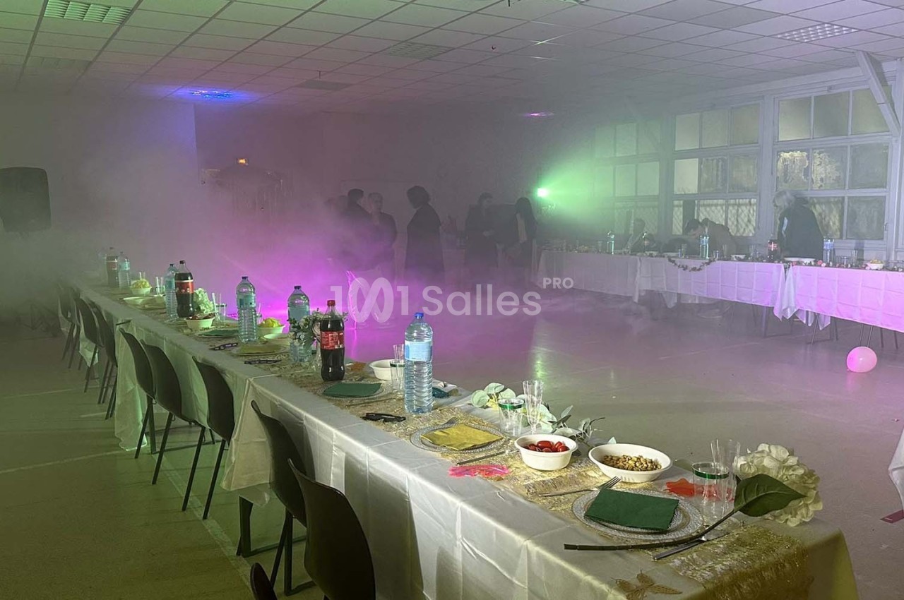 Salle de réception avec tables dressées, éclairage tamisé et fumée diffuse, quelques personnes en arrière-plan.