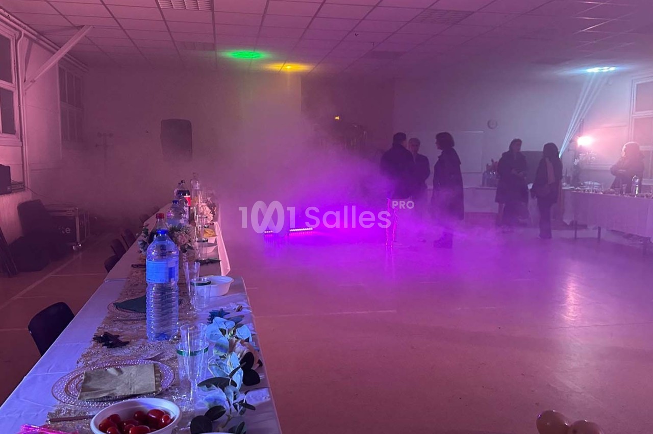 Salle de réception avec tables dressées, éclairage coloré et fumée légère, quelques personnes en arrière-plan.