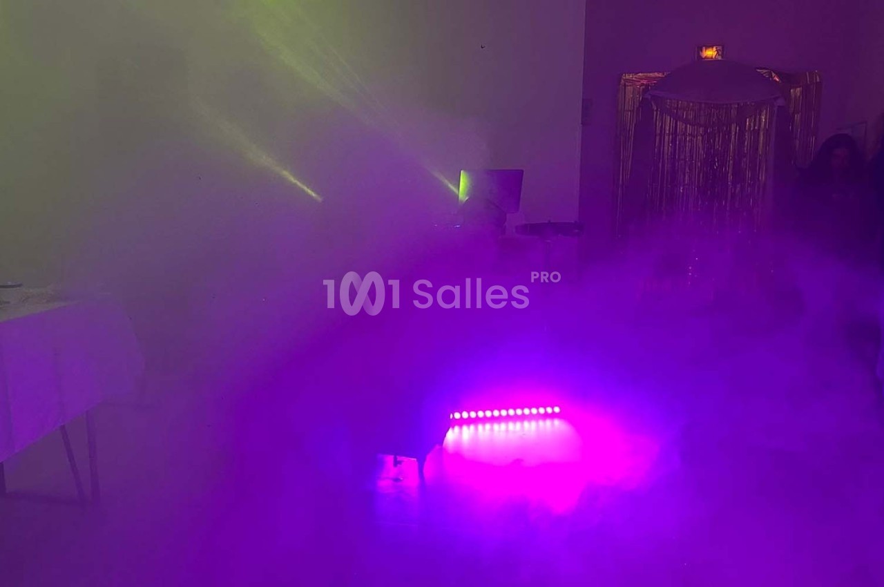 Salle obscure avec éclairage violet et vert, fumée diffuse, table dressée sur la gauche et rideaux brillants en arrière-plan.