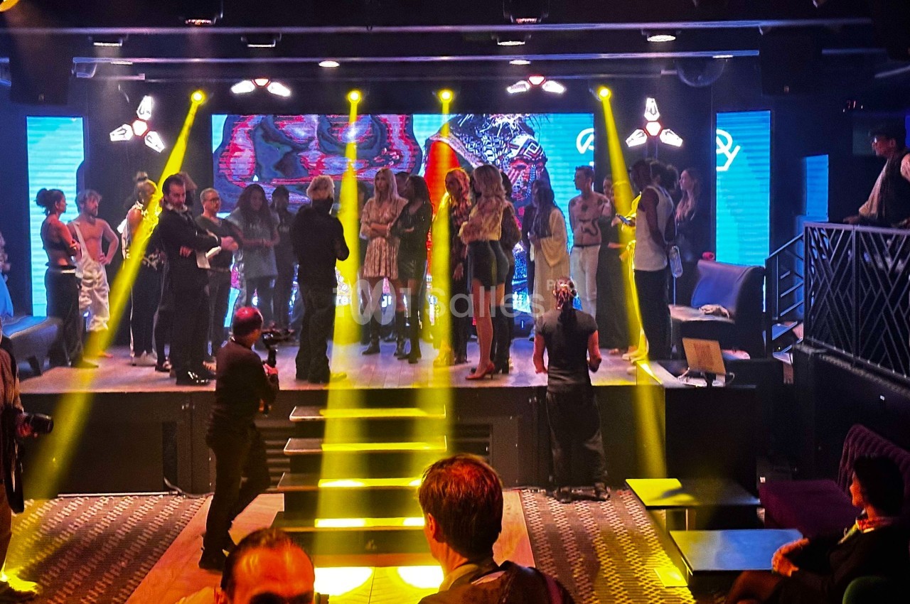 Groupe de personnes rassemblées sur une scène illuminée par des projecteurs jaunes dans un club ou une salle de spectacle.