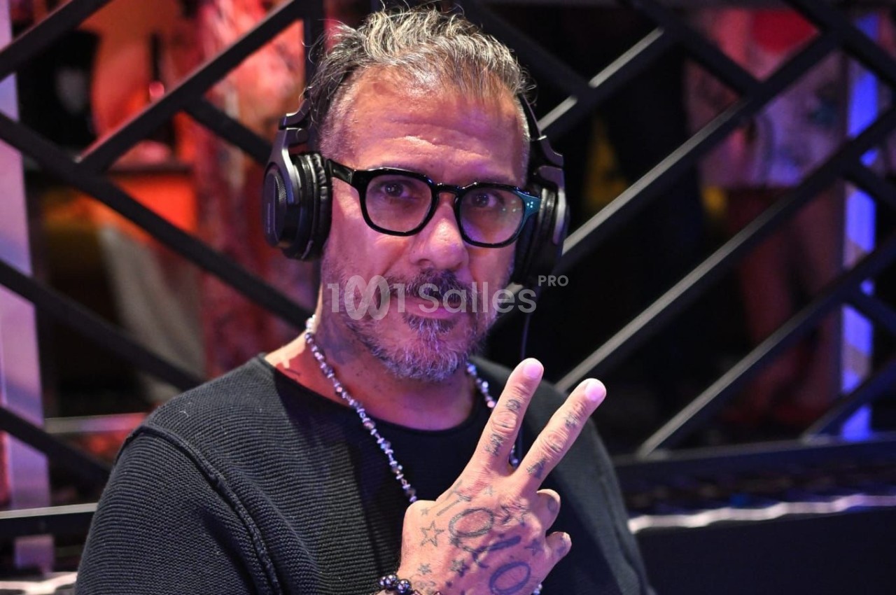 Homme portant des lunettes et un casque audio, faisant un signe de la main devant une grille métallique.