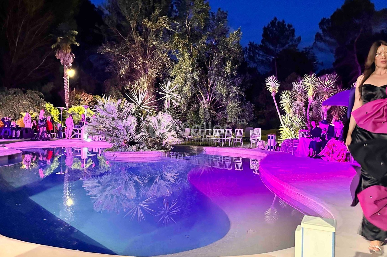 Une femme en robe noire et rose marche près d'une piscine éclairée, entourée de végétation et de convives en soirée.