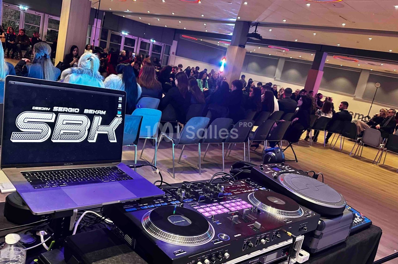 Vue d'une salle de conférence avec un DJ installé au premier plan et un public assis en arrière-plan.