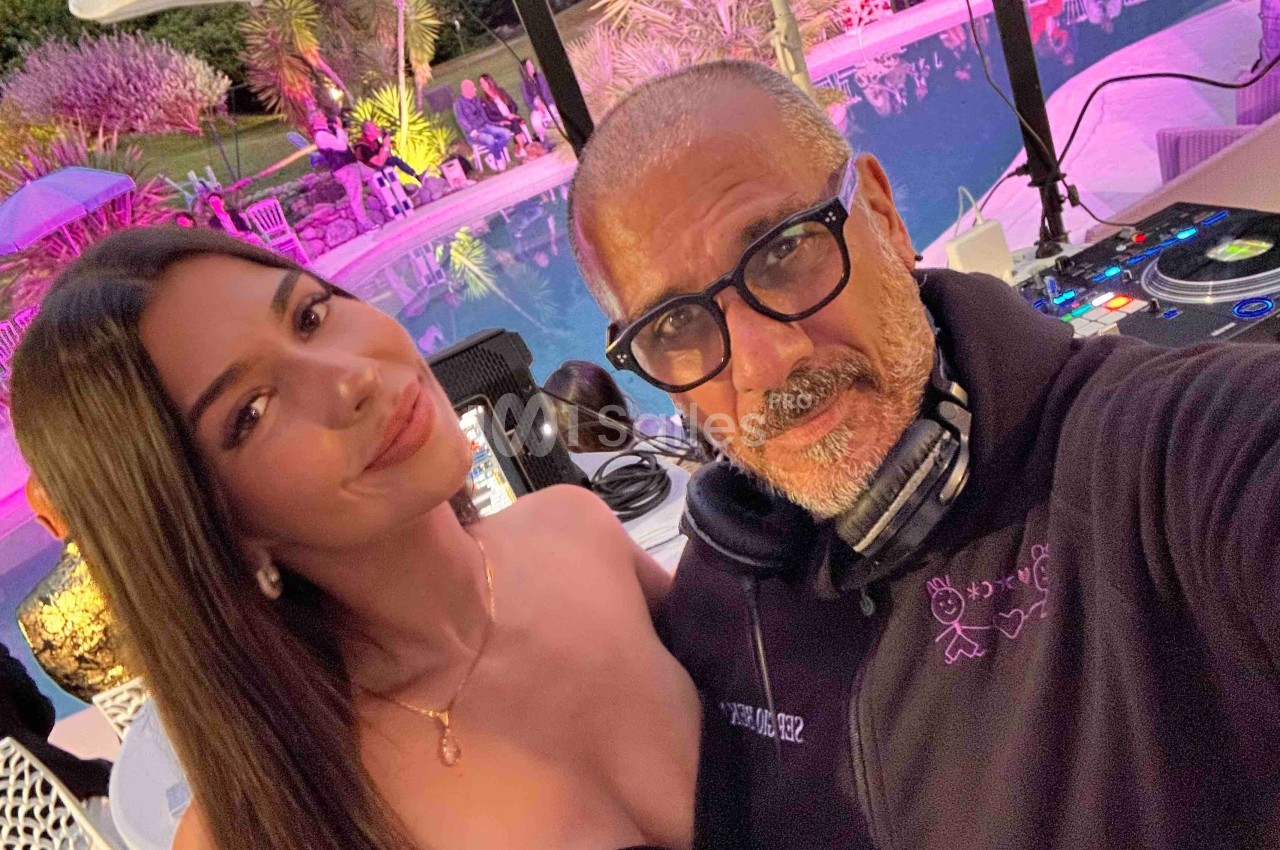 Un homme et une femme posent devant une table de mixage près d'une piscine éclairée en soirée.