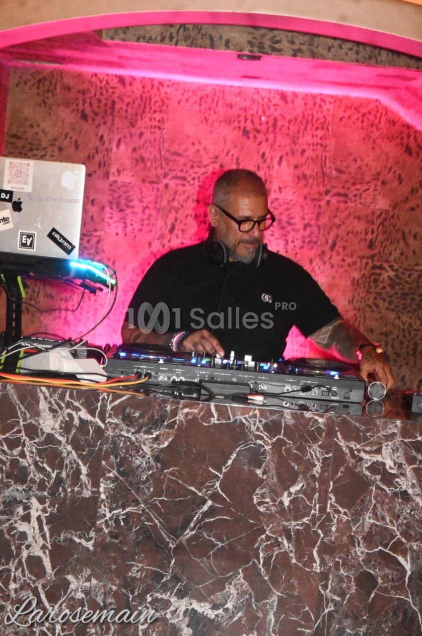 DJ en cabine équipée de platines, éclairage rose et fond texturé.