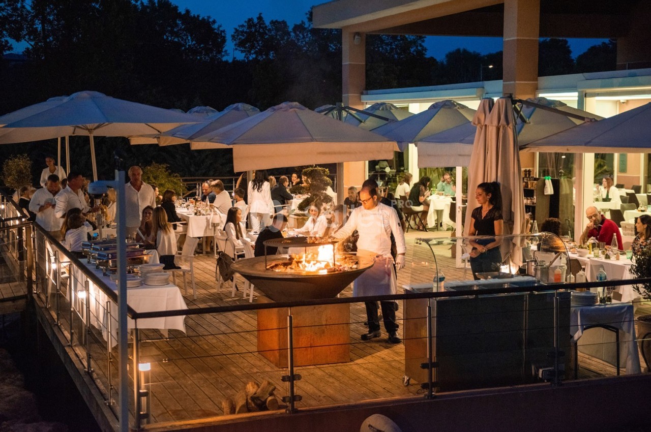 Terrasse d'un restaurant en soirée avec des convives attablés et un chef cuisinant sur un brasero.