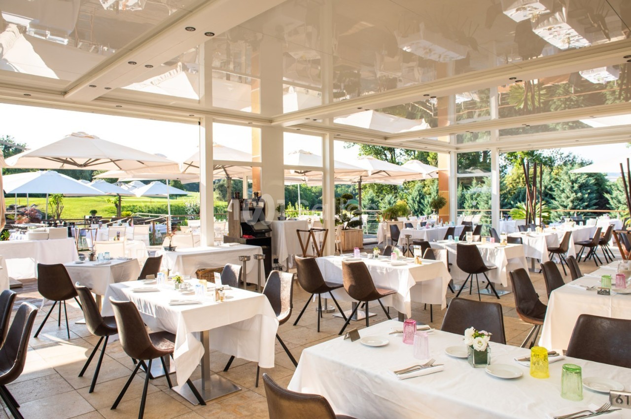 Salle de restaurant lumineuse avec grandes baies vitrées, tables dressées et terrasse extérieure avec parasols.