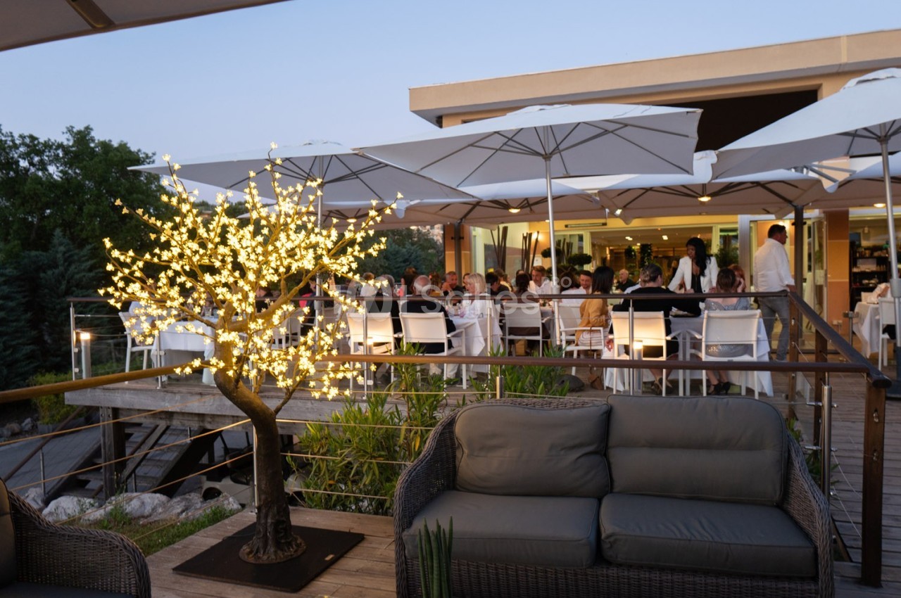 Terrasse d'un restaurant en soirée avec des convives attablés sous des parasols et un arbre lumineux décoratif au premier…
