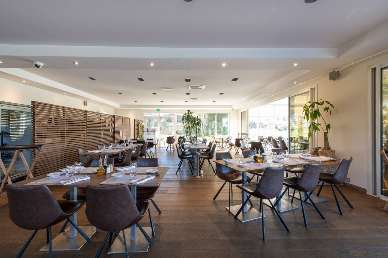 Salle de restaurant lumineuse avec tables dressées, chaises modernes et grandes baies vitrées donnant sur l'extérieur.