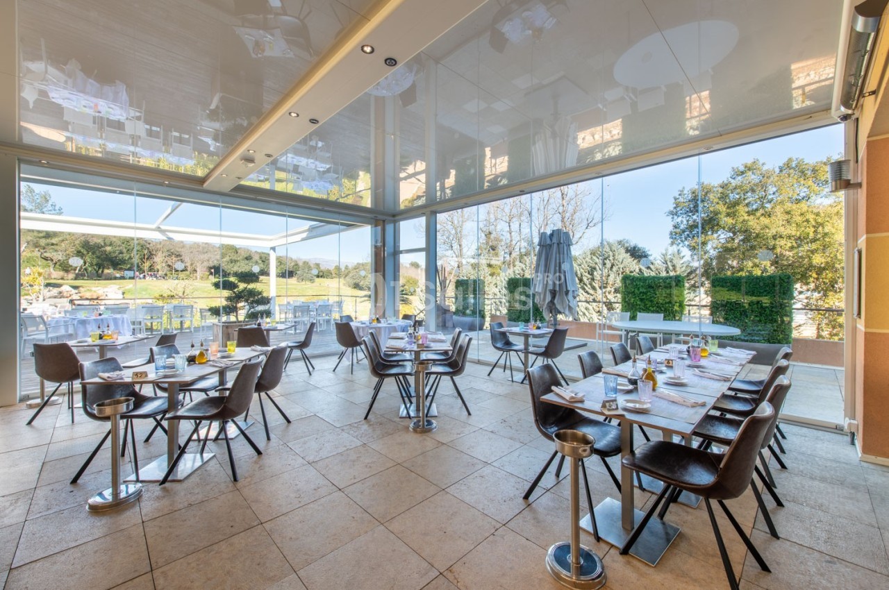 Salle de restaurant lumineuse avec grandes baies vitrées donnant sur une terrasse et un paysage verdoyant.