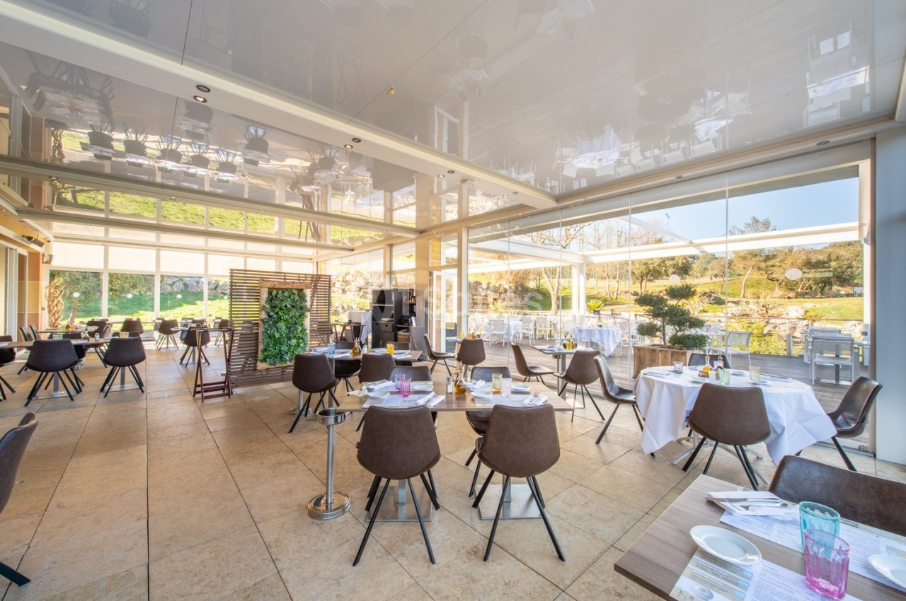 Salle de restaurant lumineuse avec baies vitrées, tables dressées et vue sur un jardin extérieur verdoyant.