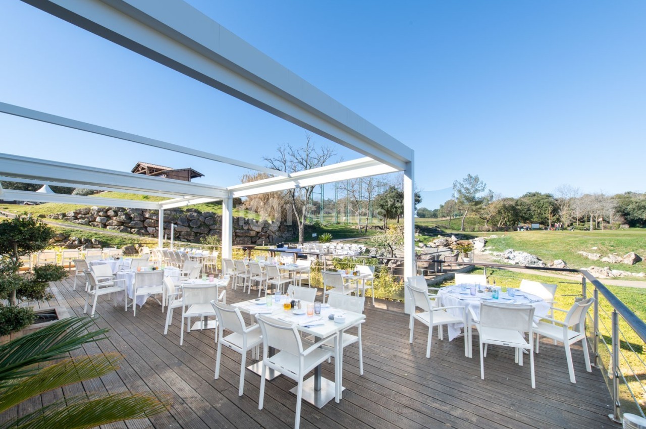 Terrasse en bois aménagée avec des tables et chaises blanches, entourée de verdure sous un ciel dégagé.