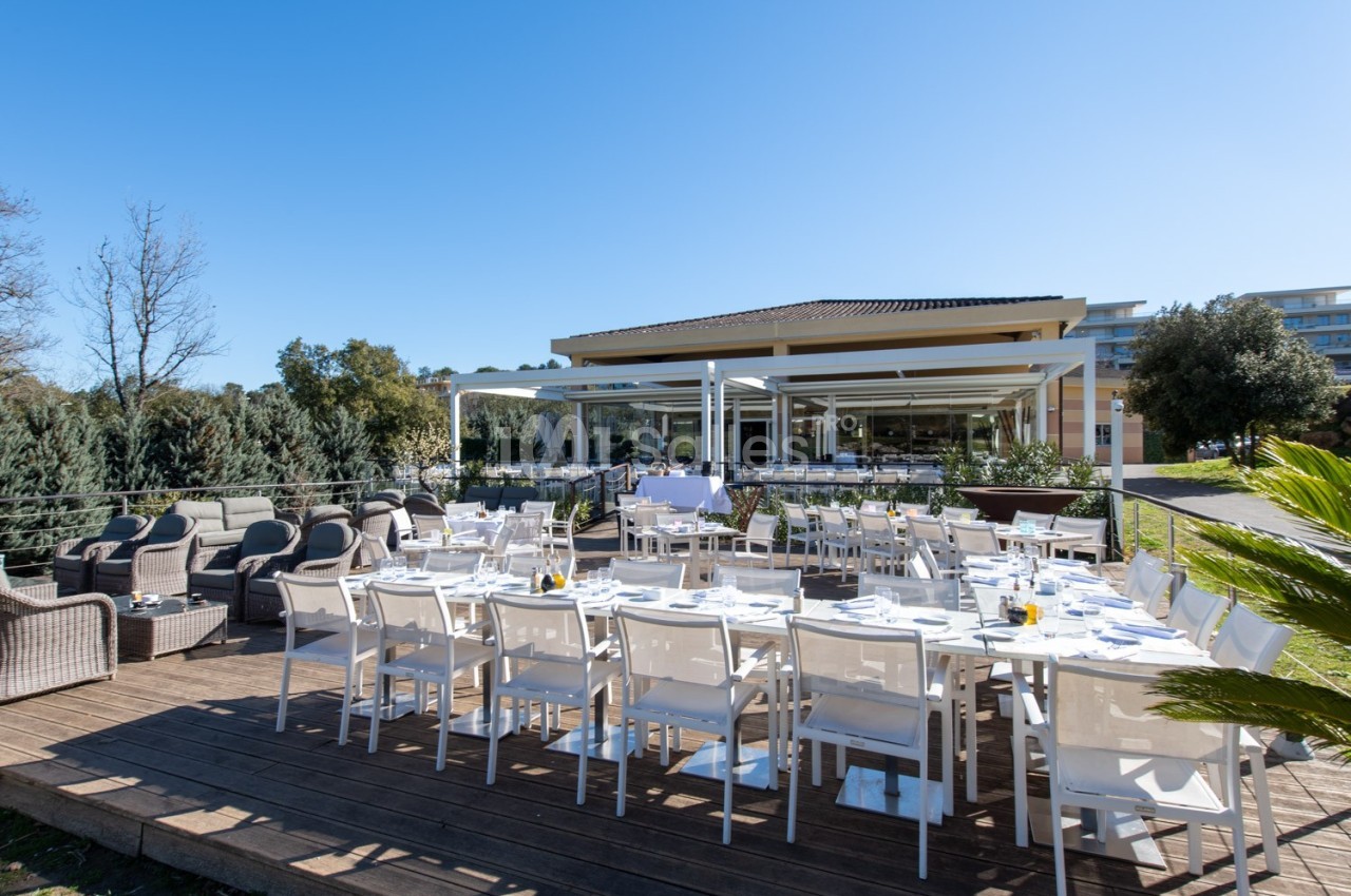 Terrasse aménagée avec tables dressées et chaises blanches, entourée de végétation et donnant sur un bâtiment moderne.