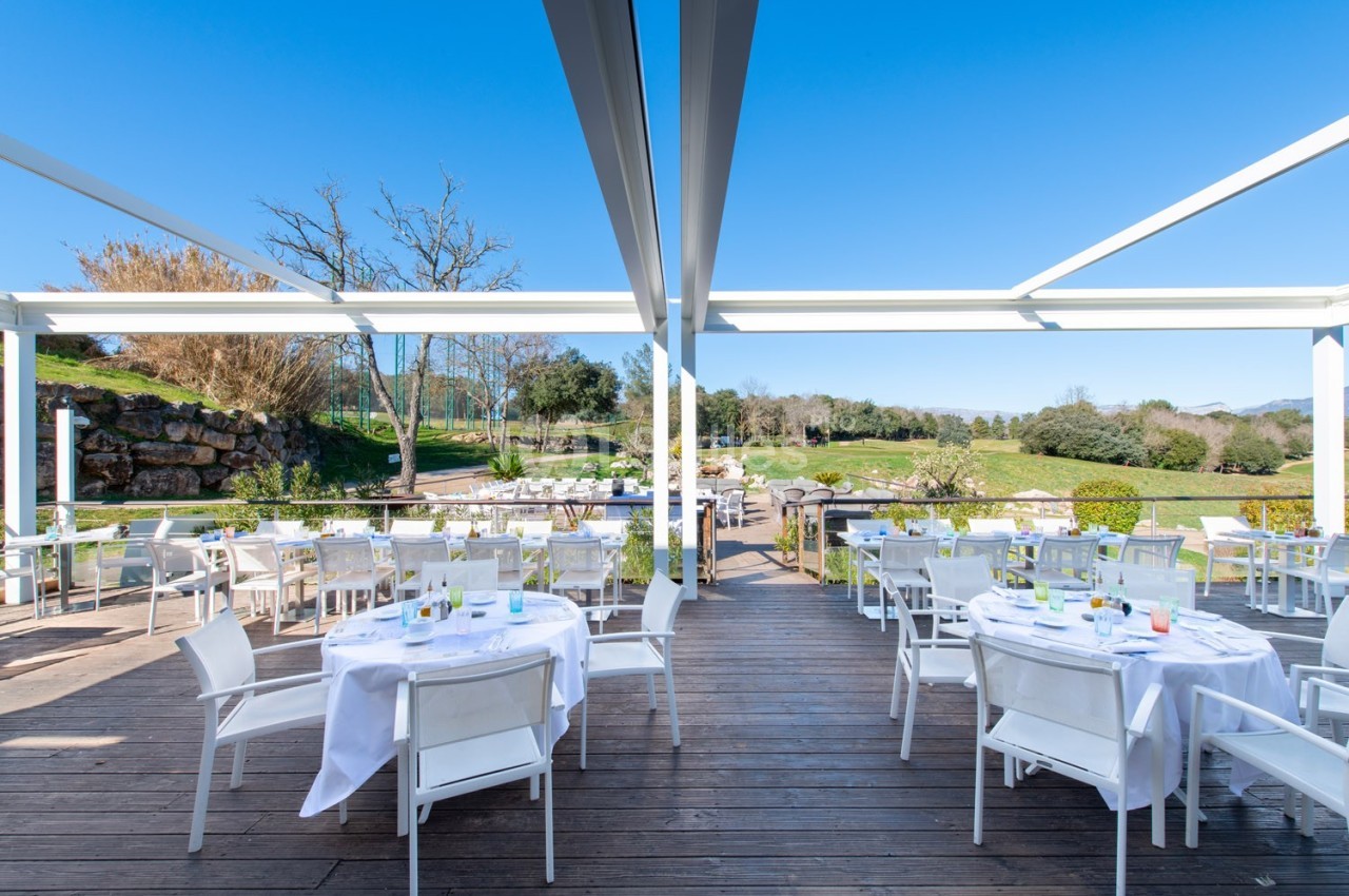 Terrasse en bois avec tables dressées sous une pergola, offrant une vue sur un jardin verdoyant par temps ensoleillé.