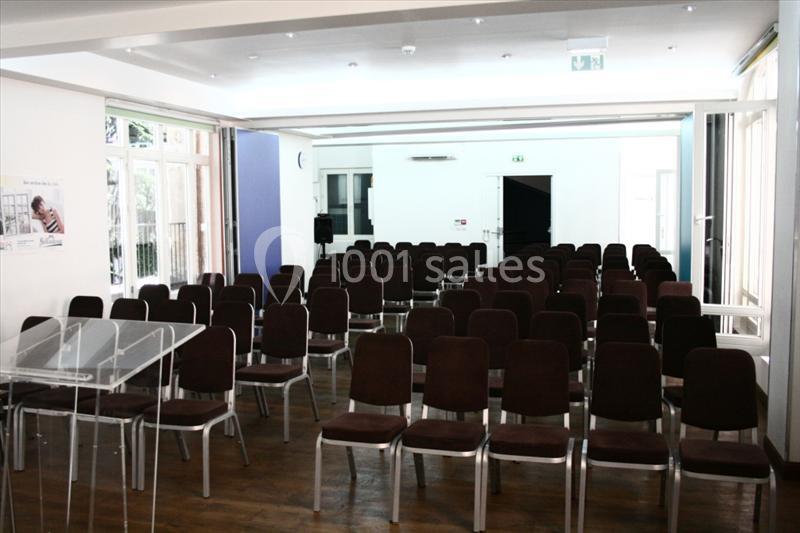 Salle de conférence lumineuse avec des rangées de chaises vides, un pupitre transparent et des portes ouvertes.
