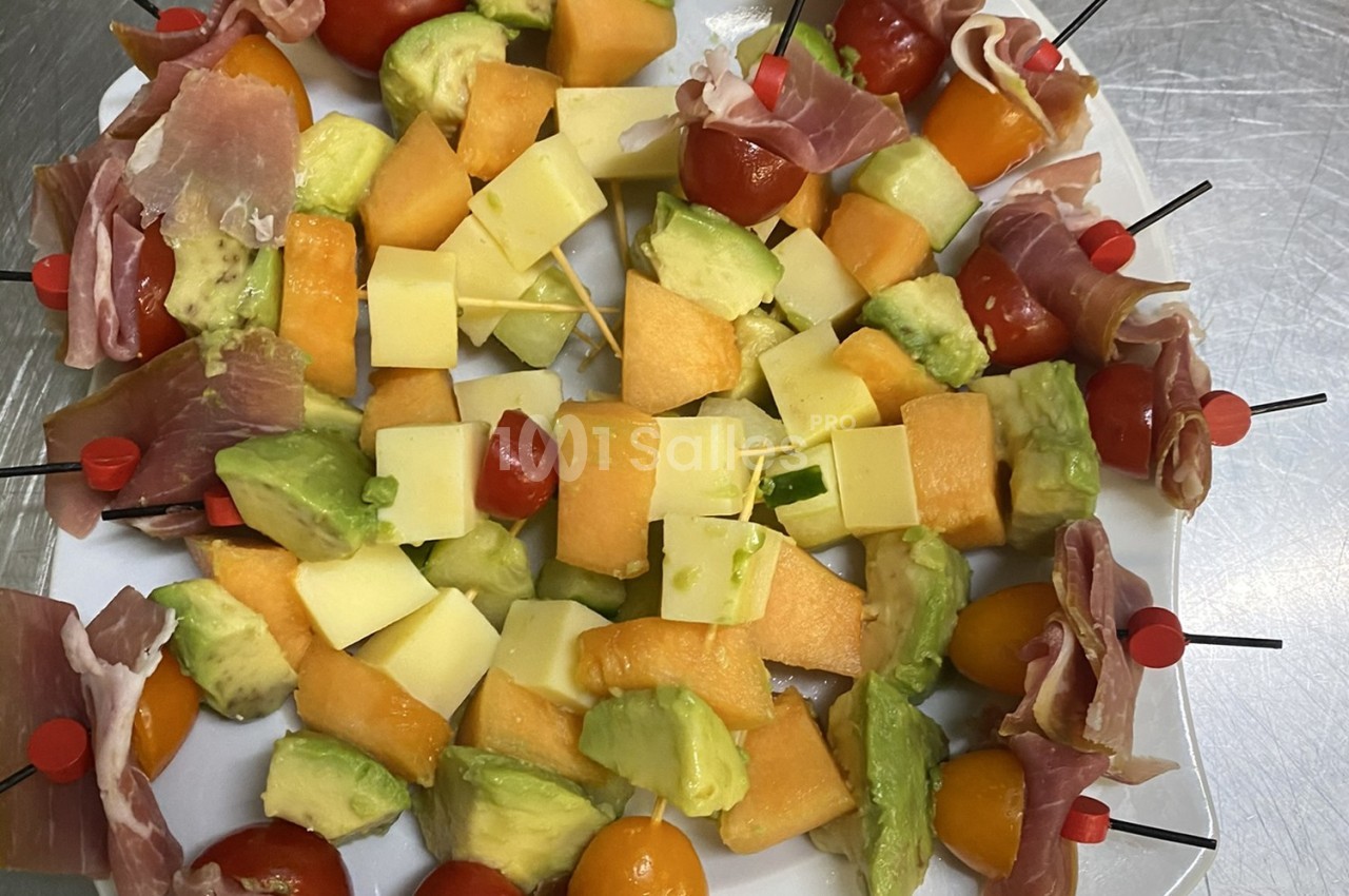 Assortiment de brochettes apéritives avec melon, avocat, fromage, tomates cerises et jambon cru sur un plateau.