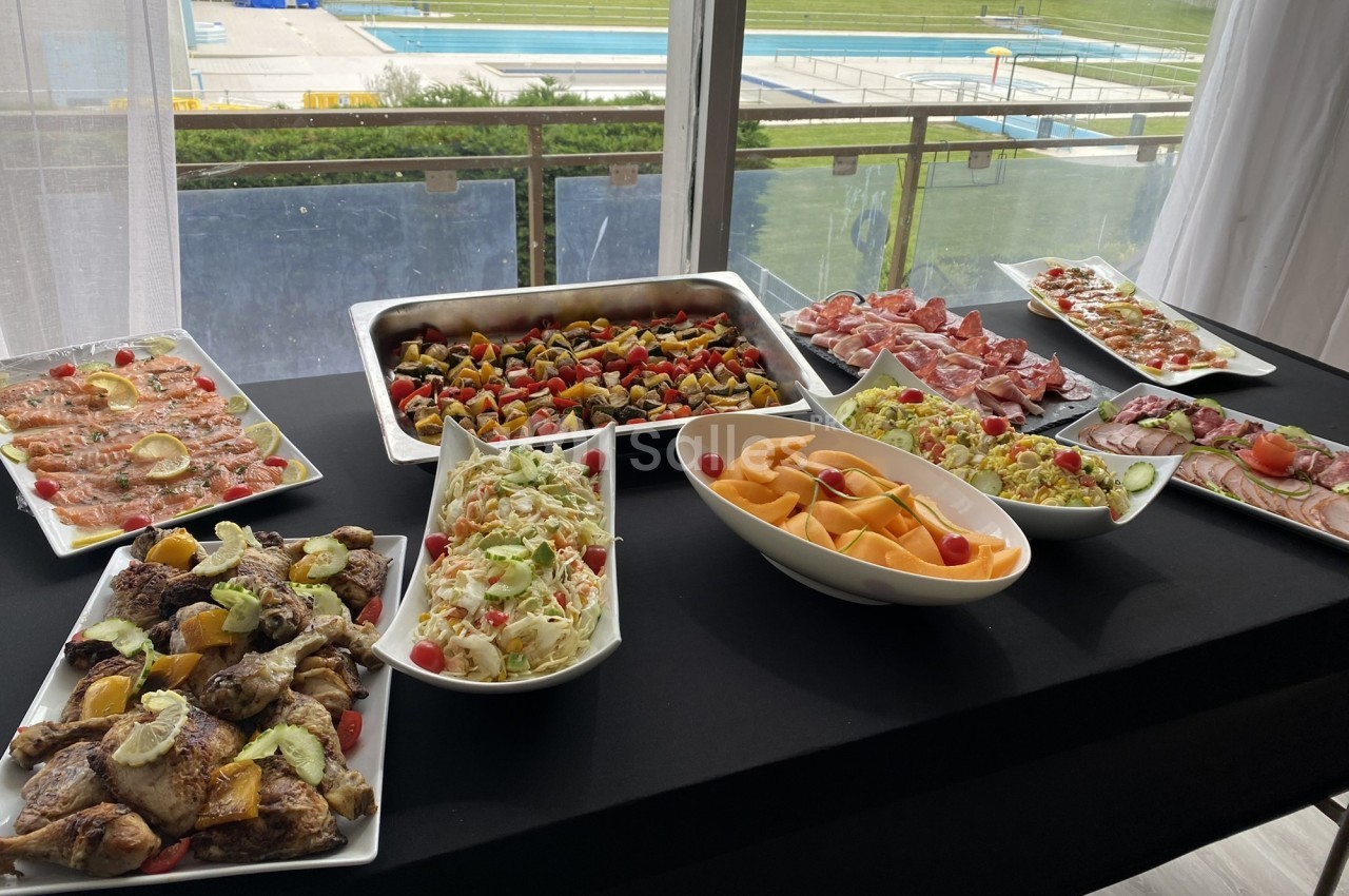 Buffet varié avec plats de viande, salades, fruits et légumes, disposé sur une table près d'une piscine extérieure.