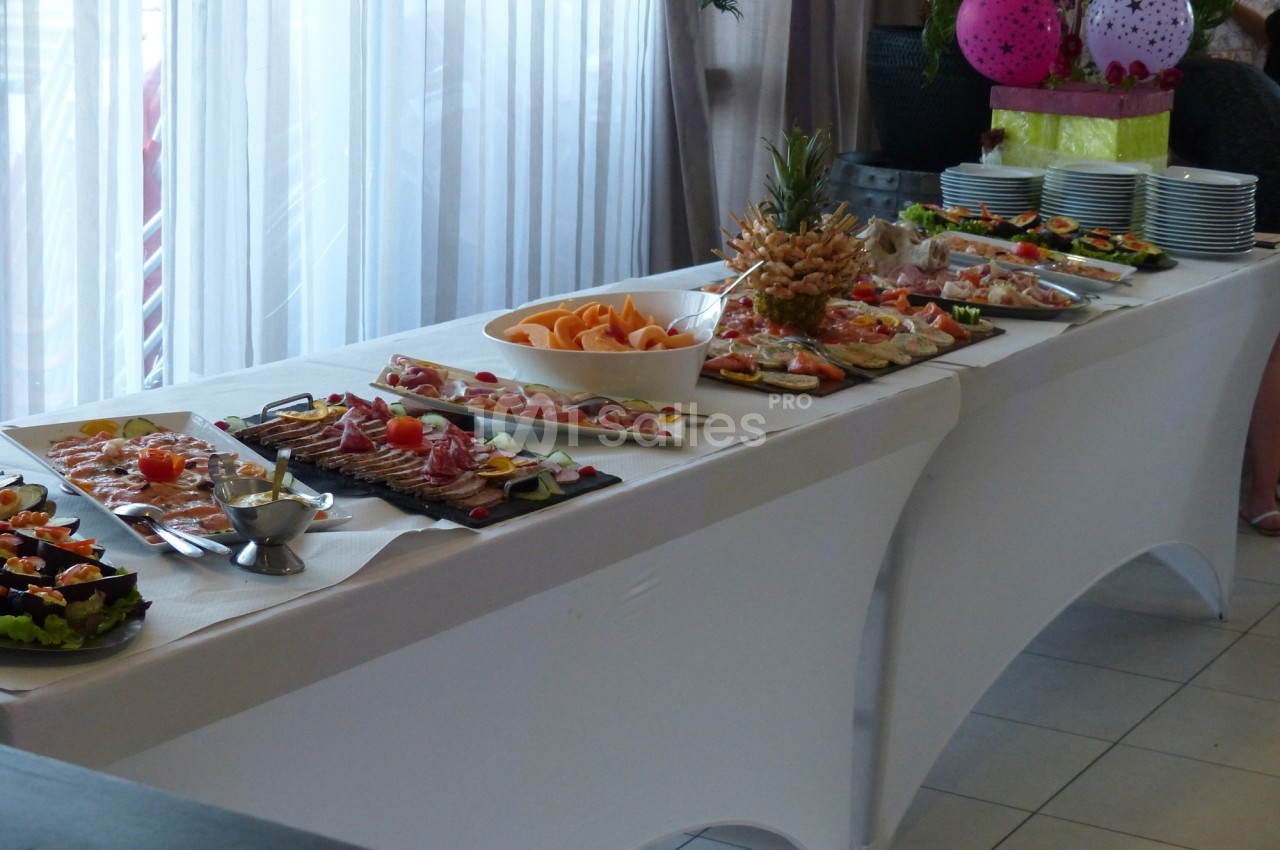 Buffet avec divers plats froids, fruits et assiettes disposés sur une table blanche près d'une fenêtre.
