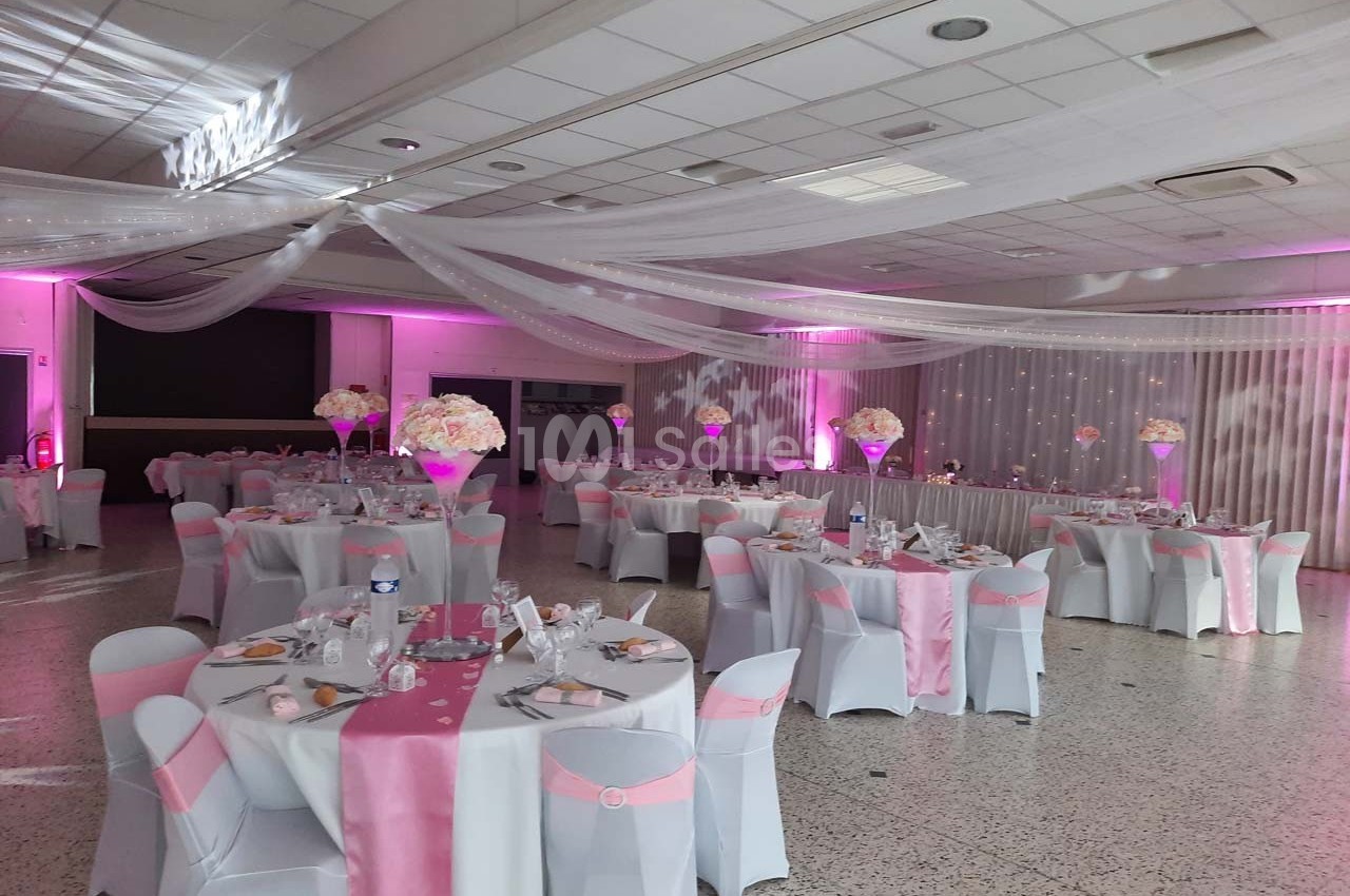 Salle de réception décorée avec des nappes blanches et roses, des centres de table fleuris et un éclairage rose.